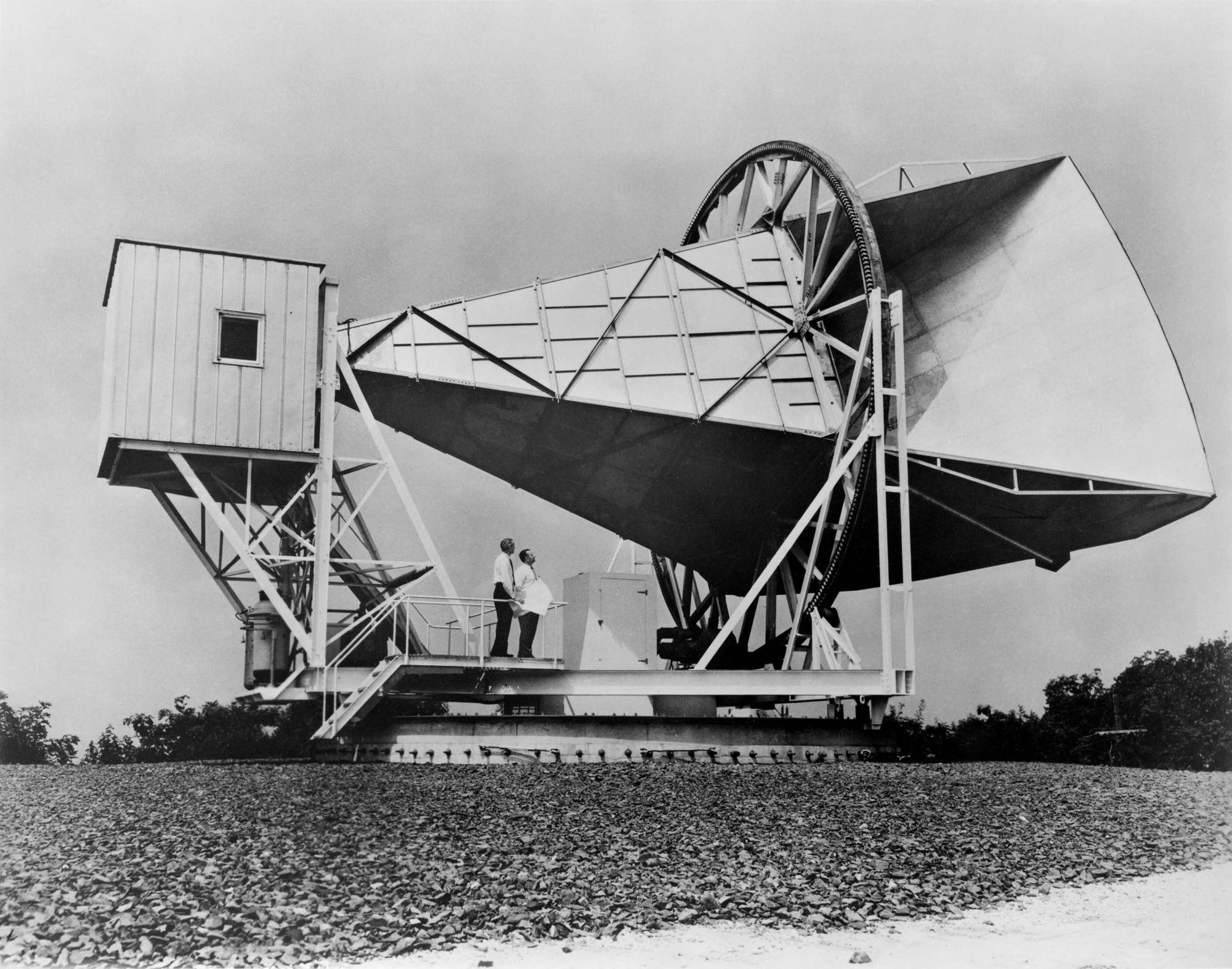 A large horn antenna that's appears on its side with two men standing beneath it. Visually the antenna looks to be about 3 men high and 10 men long. It's a Black and White photo.